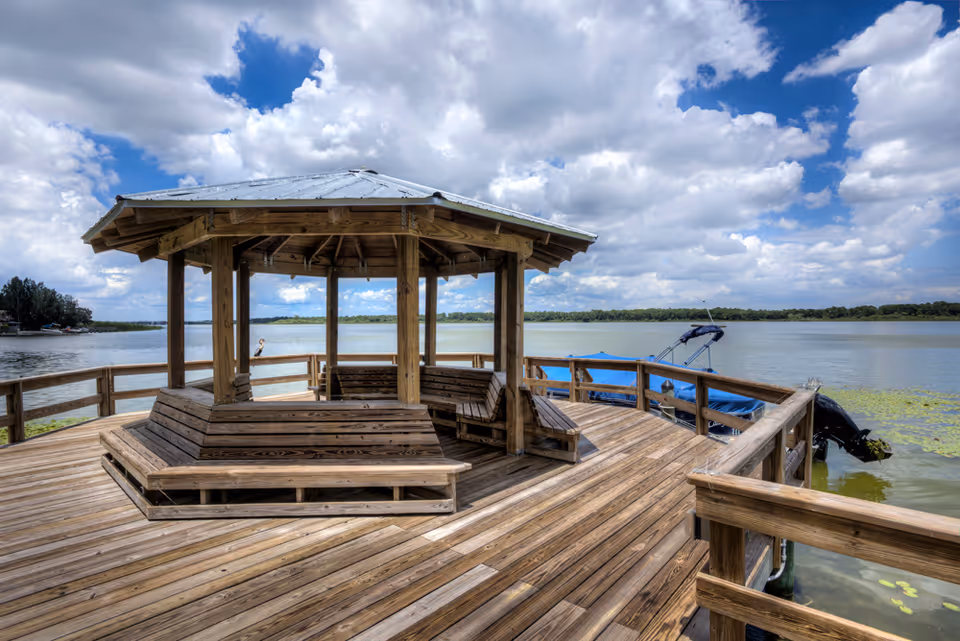 A wooden gazebo with built-in benches on a large wooden dock overlooking a calm lake under a partly cloudy sky. A boat with a blue cover is moored to the dock on the right side.