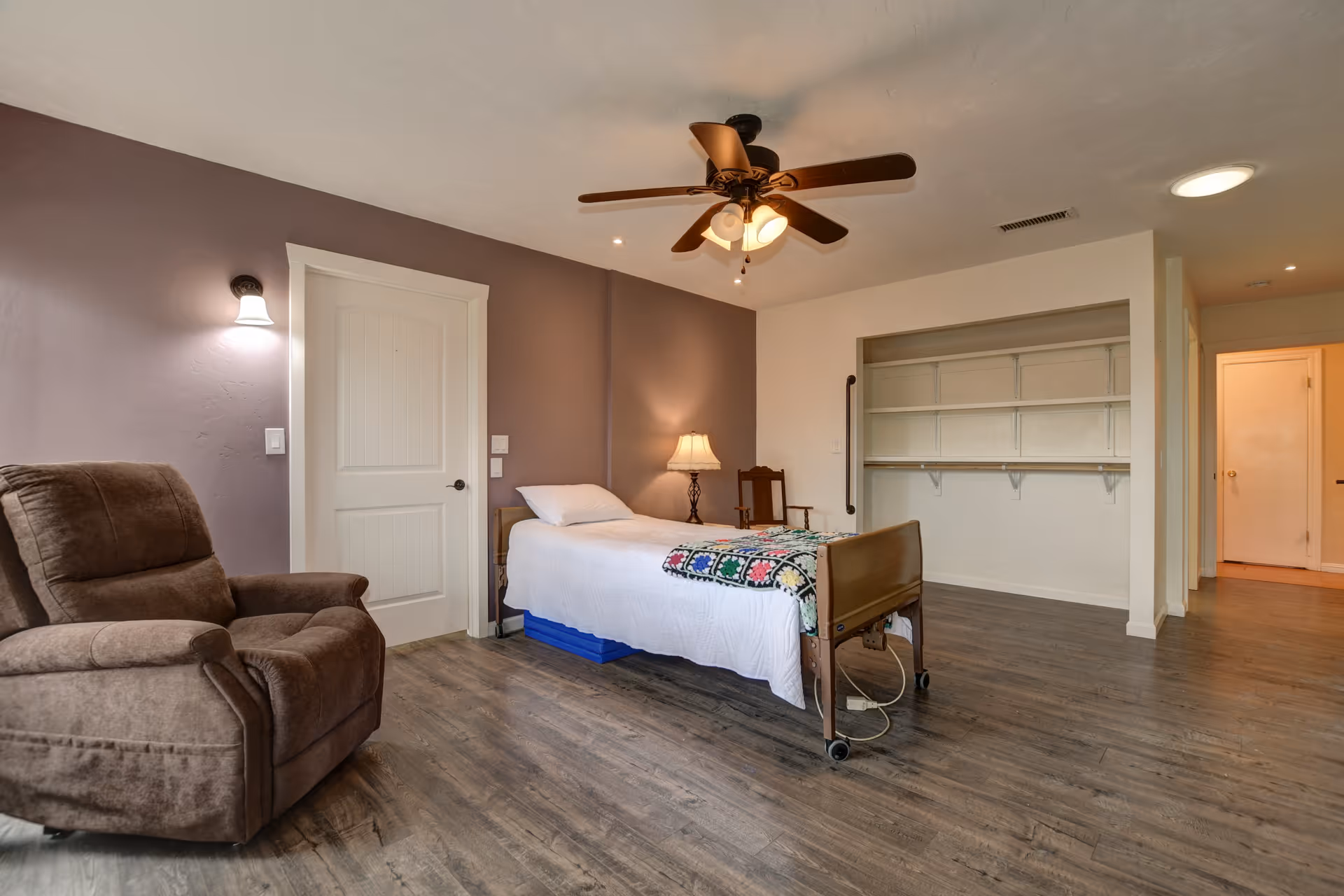 A spacious assisted living bedroom with a single bed covered in white bedding and a colorful crocheted blanket. There is a brown recliner chair to the left, a wooden chair with a lamp on a small table beside the bed, and an open closet with shelves on the right. The room has wood flooring, a ceiling fan with lights, and walls painted in light beige and muted purple tones.