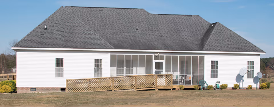 Exterior view of a single-story white building with a gray shingled roof, featuring a wooden ramp leading to a screened porch area. There are several windows, two satellite dishes on the right side, and some bushes around the building. The sky is clear and blue.