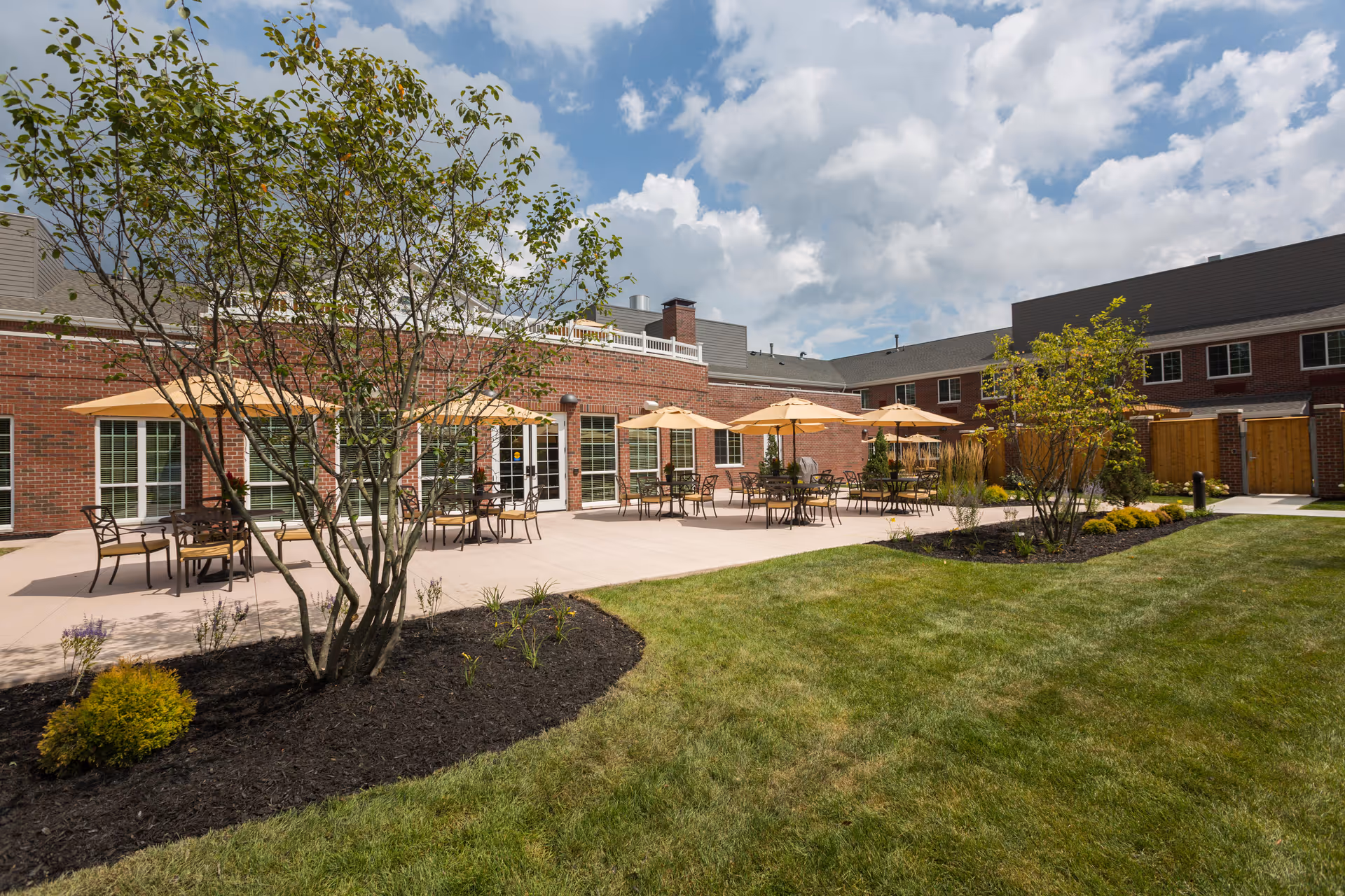 Outdoor patio area at StoryPoint Gahanna North with multiple tables and chairs under beige umbrellas, surrounded by landscaped garden beds and green grass, adjacent to a brick building under a partly cloudy sky.