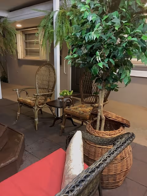 Outdoor patio area with wicker chairs and a small table. There is a potted plant with green leaves in a woven basket, and hanging ferns are visible near a window and door in the background.