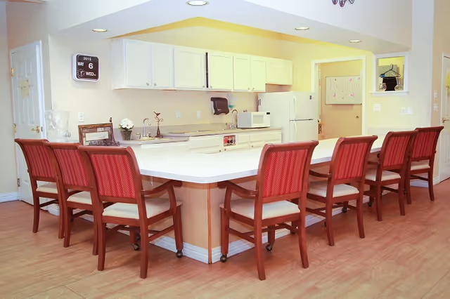 A bright kitchen area with a large white countertop island surrounded by seven wooden chairs with red cushions. The kitchen has white cabinets, a microwave, a refrigerator, and a paper towel dispenser. The floor is wooden, and there is a wall clock showing the date May 6, 2015.