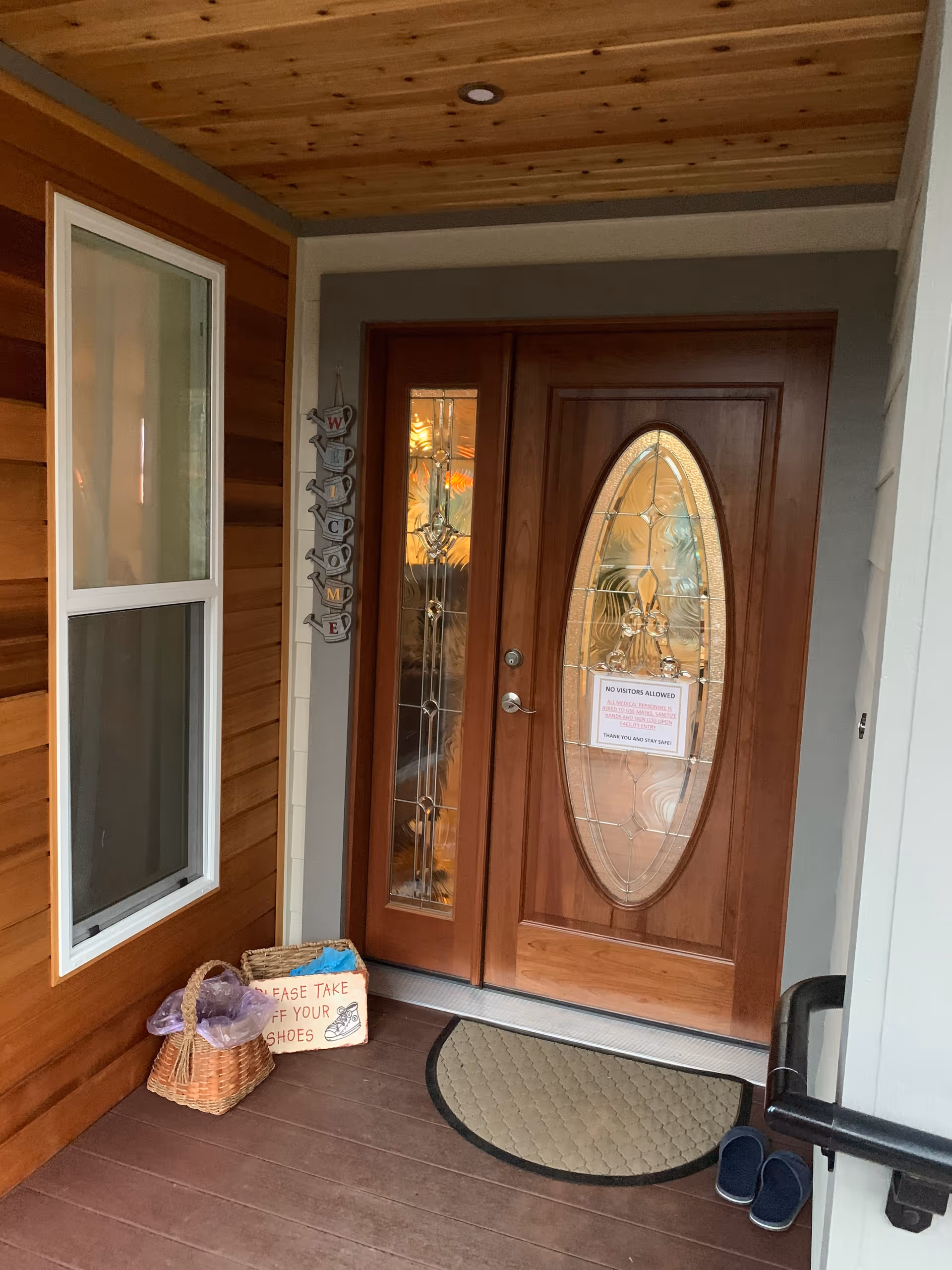 Front entrance of a facility with double wooden doors featuring decorative glass panels. A welcome sign made of watering cans hangs on the wall next to the door. There is a basket with a sign that says 'Please take off your shoes' and a pair of slippers on the floor near a small mat. The walls are wooden and there is a window to the left of the door.