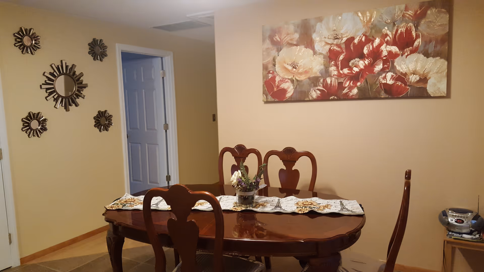A dining room with a polished wooden table and four matching chairs. The table has a decorative runner and a small floral centerpiece. On the wall above the table is a large painting of red and white flowers. To the left, there are five decorative round mirrors arranged on a beige wall. A doorway with a white door is visible in the background, and a small stereo system sits on a wooden side table to the right.