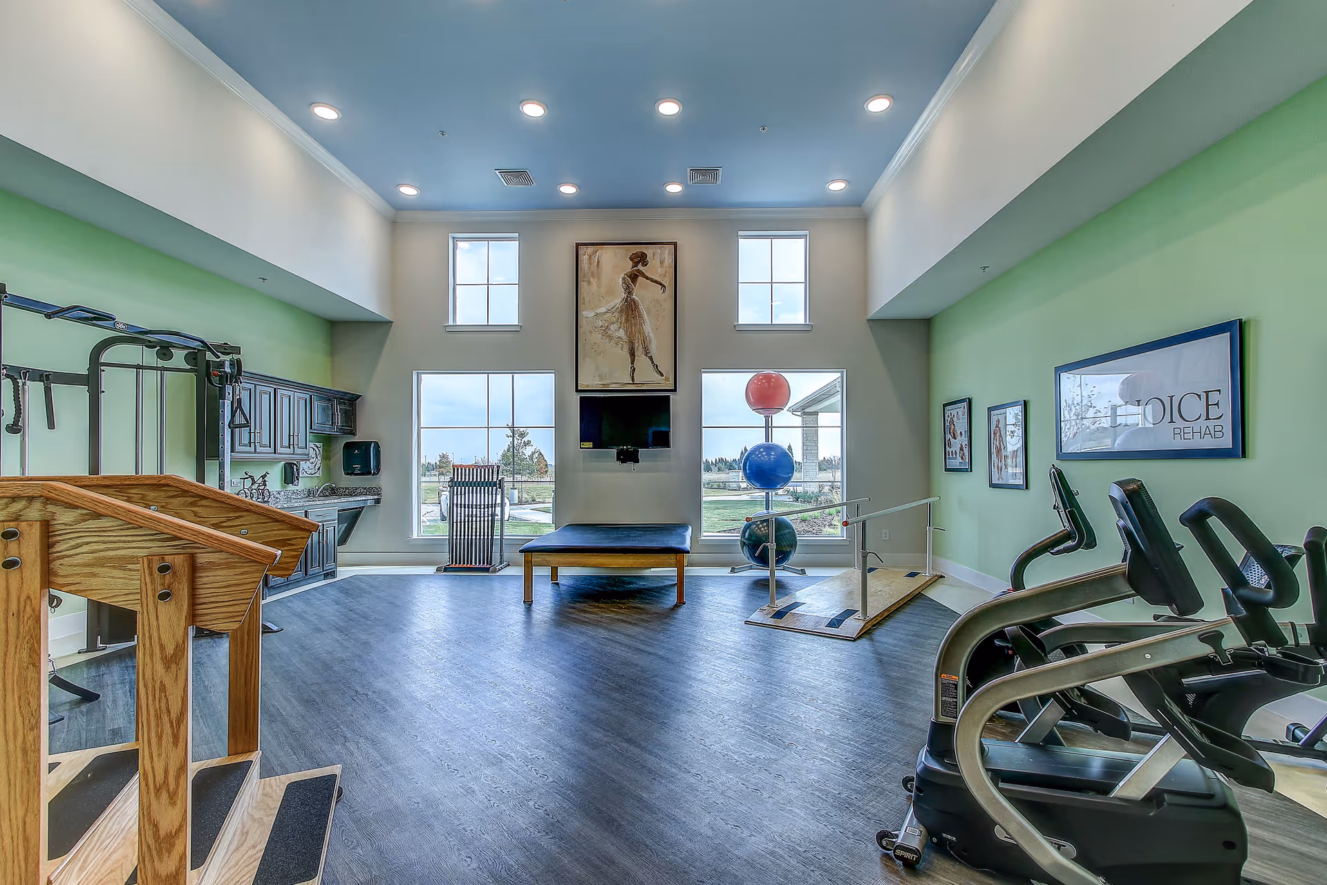 A bright rehabilitation exercise room with large windows letting in natural light. The room features exercise equipment including stationary bikes, a wooden ramp with handrails, a set of colorful exercise balls stacked on a stand, and a wooden therapy table. The walls are painted light green and decorated with framed pictures and a large painting of a ballerina. There is a sink and cabinets on the left side of the room.