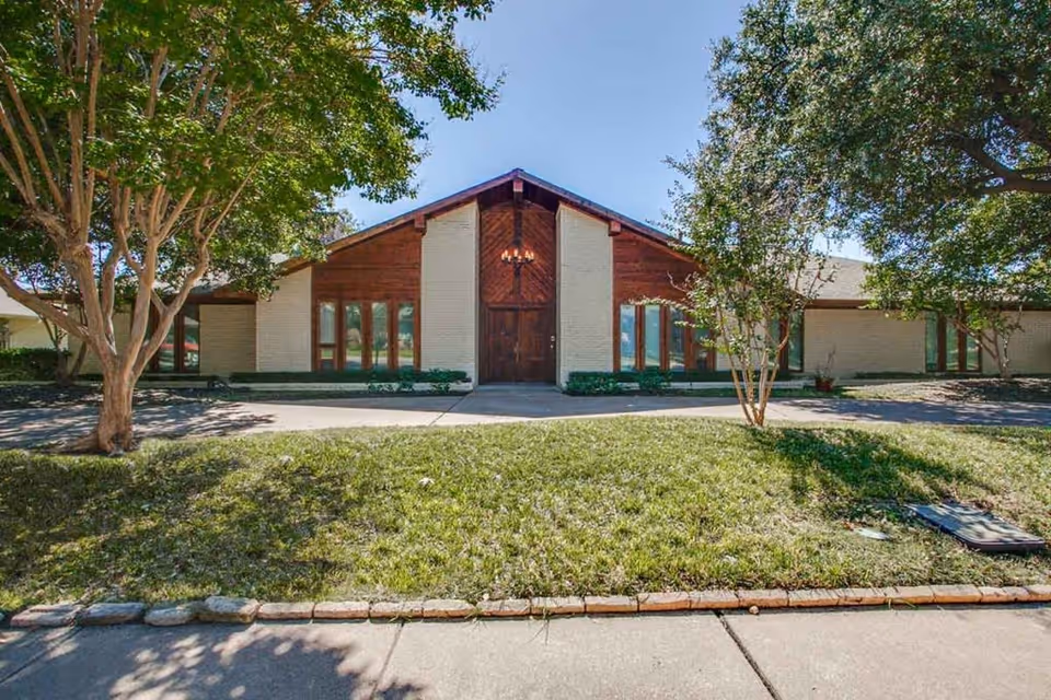 Front exterior view of a single-story building with a peaked roof, large wooden double doors, and tall windows on either side. The building is surrounded by green grass, trees, and a sidewalk in front under a clear blue sky.