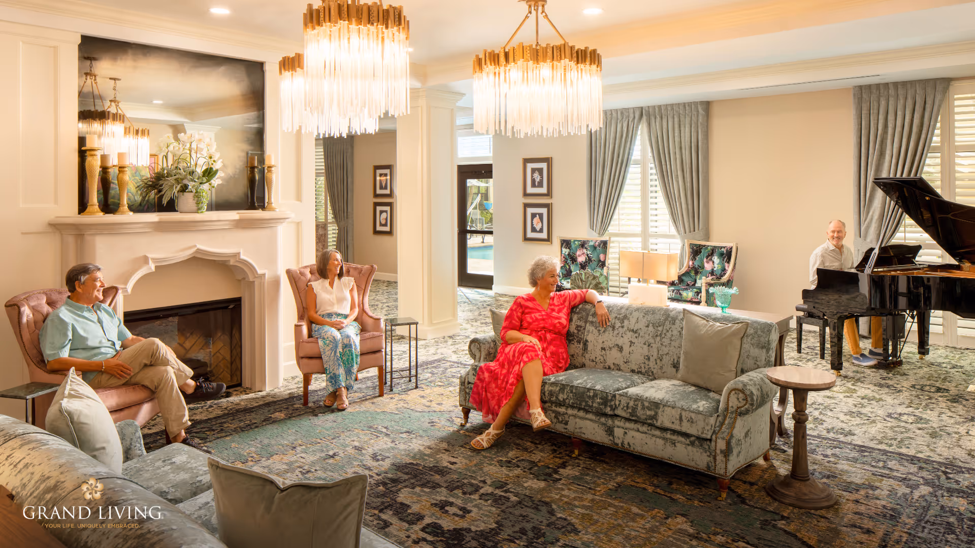 A bright and elegant living room in a senior living facility with three elderly people seated on plush chairs and a sofa, and one elderly man playing a grand piano. The room features a fireplace with decorative vases and flowers, large windows with curtains, two chandeliers, and a patterned carpet.