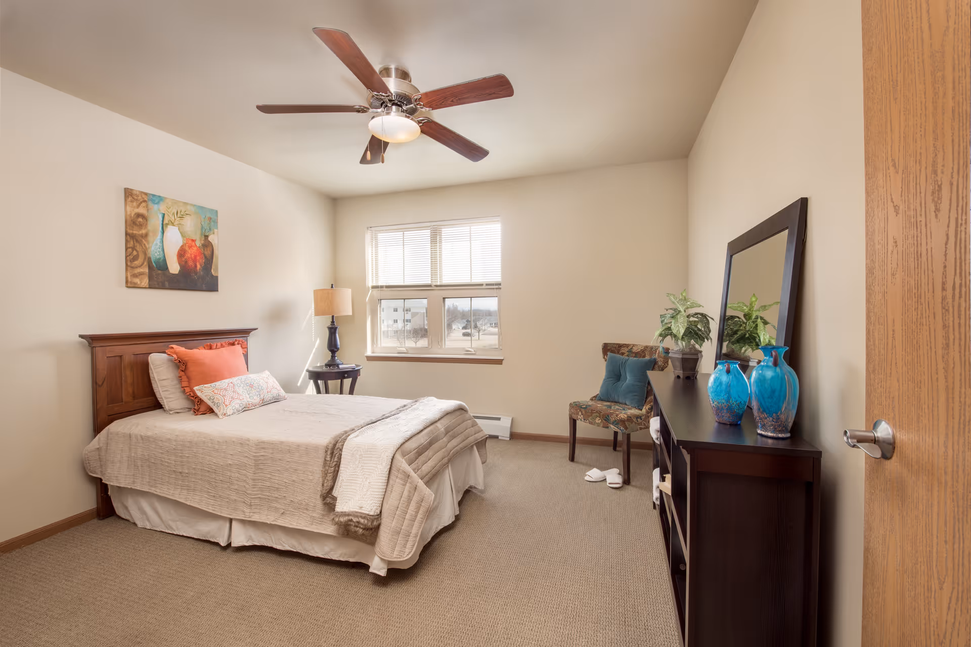 A neatly arranged bedroom with a bed featuring beige bedding and decorative pillows, a wooden headboard, a nightstand with a lamp, a ceiling fan, a window letting in natural light, a patterned chair with a teal cushion, a dark wooden dresser with a large mirror, blue vases, and a potted plant.
