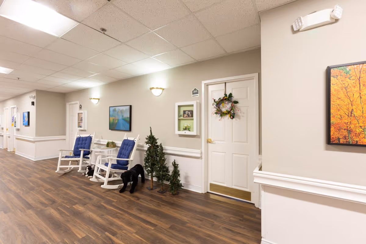 A well-lit hallway in a senior living facility with wooden flooring, beige walls, and white doors. Two white rocking chairs with blue cushions are placed next to a small white table. There are small decorative trees and a black dog statue near one door, which has a wreath hanging on it. Paintings and wall-mounted lights decorate the walls.