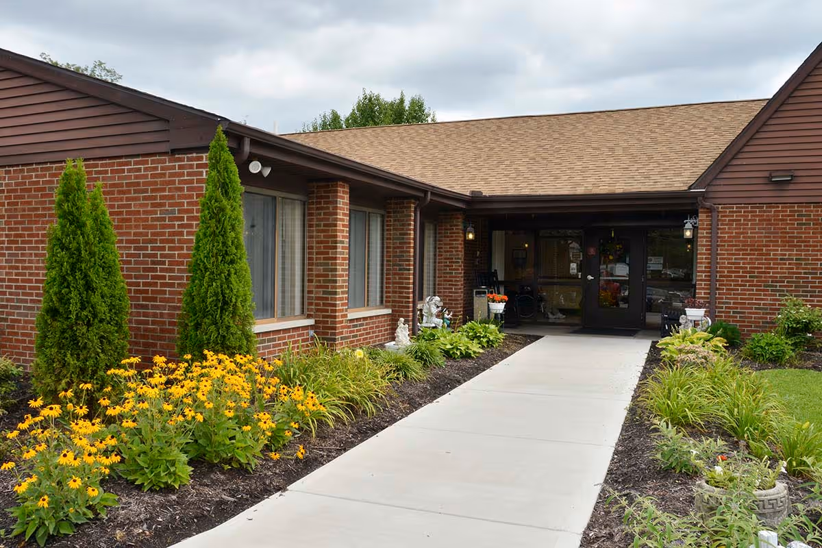 Exterior view of Wayne Healthcare Center showing a brick building with a sloped roof, a concrete walkway leading to the entrance, and landscaped flower beds with yellow flowers and green shrubs on either side.