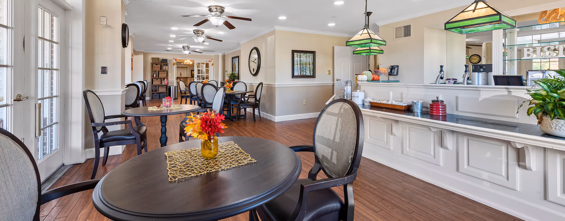 A bright and spacious dining area in a senior living facility with several round tables and chairs. The tables have decorative centerpieces with flowers. The room features wooden flooring, ceiling fans, and large windows letting in natural light. A counter area with green pendant lights and a coffee station is visible on the right side.