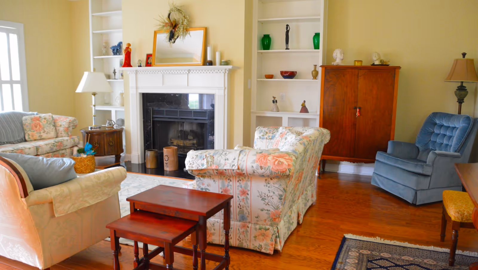 A cozy living room with floral patterned sofas, a blue armchair, a wooden cabinet, and a white fireplace with a mirror and decorative items on the mantel. The room has wooden floors, built-in white shelves with various ornaments, and two table lamps.