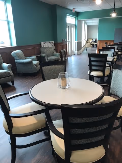 A spacious common area with round tables and cushioned chairs arranged for seating. The room has teal walls, large windows letting in natural light, and several armchairs along the wall. The flooring is dark wood, and there is a glass candle holder on the nearest table.