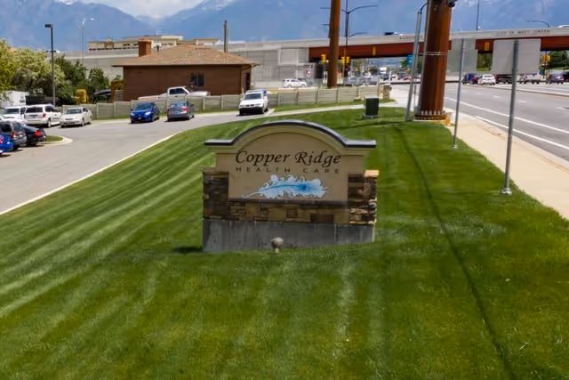 Stone sign reading 'Copper Ridge Health Care' on a manicured grassy lawn beside a road and parking lot with cars and mountains in the background.