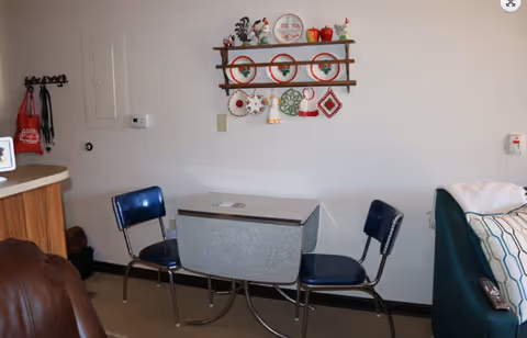 A small dining area with a vintage drop-leaf table and two blue chairs. On the wall above the table is a wooden rack displaying decorative plates and hanging kitchen towels. To the right is a green couch with a patterned pillow and a remote control on the armrest. To the left, part of a brown leather chair and a counter with a red bag hanging on a hook are visible.