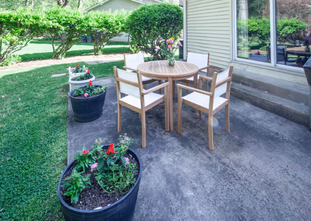 Outdoor patio area with a round wooden table and four wooden chairs with white cushions. The patio is surrounded by green grass, bushes, and several large flower pots with blooming flowers. The side of a light-colored building with a window is visible in the background.