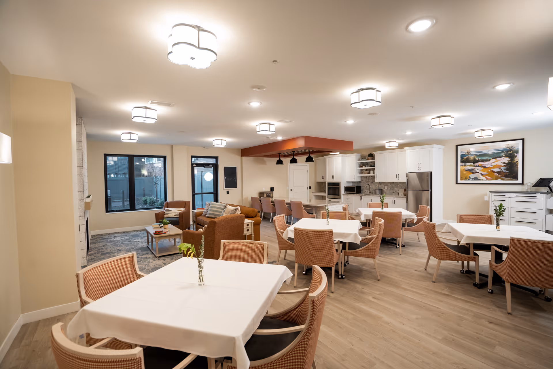 A spacious and well-lit common area in The Pillars of Lakeville featuring multiple dining tables with white tablecloths and chairs, a cozy seating area with armchairs and a sofa, and a modern kitchen with white cabinetry and stainless steel appliances. The room has light wood flooring, several ceiling lights, and a large window showing an outdoor view.
