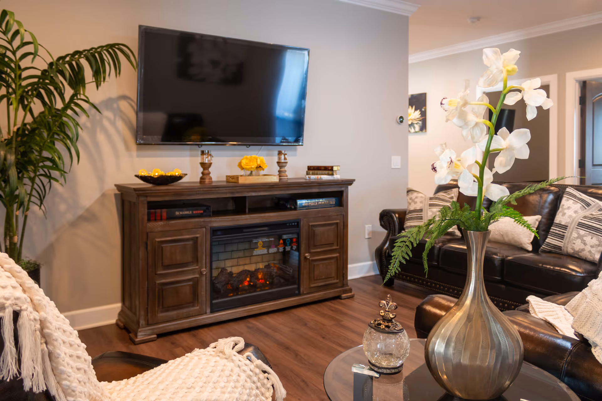 A cozy living room with a wall-mounted flat-screen TV above a wooden cabinet with an electric fireplace. The room features a dark leather sofa with patterned cushions, a round glass coffee table with a large silver vase holding white flowers and greenery, and a cream-colored knitted throw draped over a chair. There is a tall green plant in the corner and warm wooden flooring.