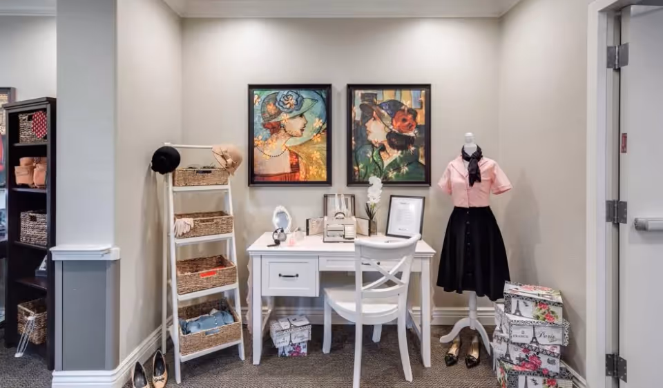 A decorated interior vignette featuring a white desk and chair, a ladder shelf with woven baskets, a mannequin dressed in vintage clothing, framed portraits on the wall, and stacked decorative boxes.