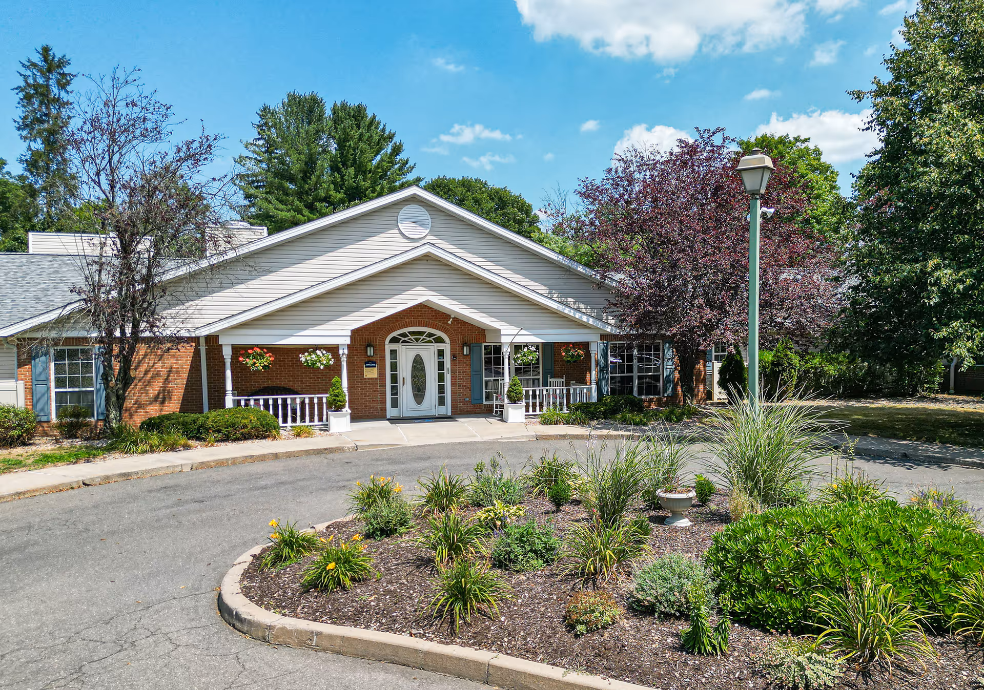 Front exterior view of a single-story brick and siding building with a covered entrance, white double doors with glass panels, hanging flower baskets, and a landscaped circular driveway with various green shrubs and plants under a blue sky with scattered clouds.