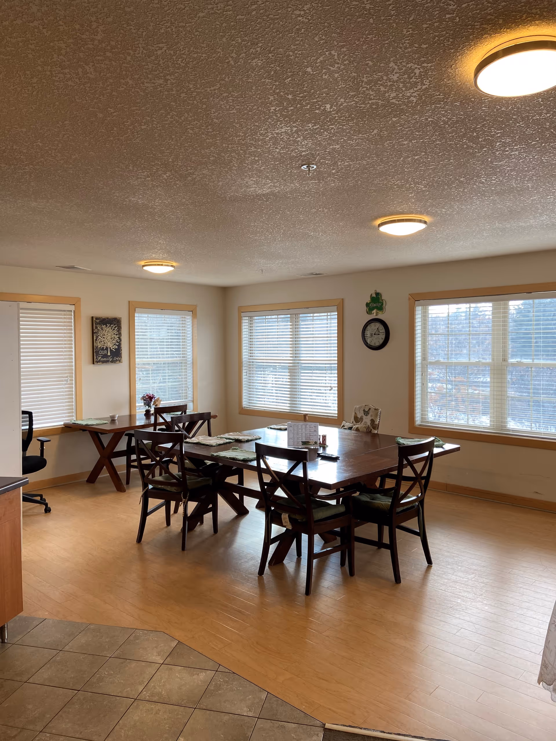 A dining room with a wooden table surrounded by six chairs with green cushions. The room has three large windows with blinds, a clock on the wall, and a small table with a chair in the corner. The floor is a combination of wood and tile, and the ceiling has three round light fixtures.