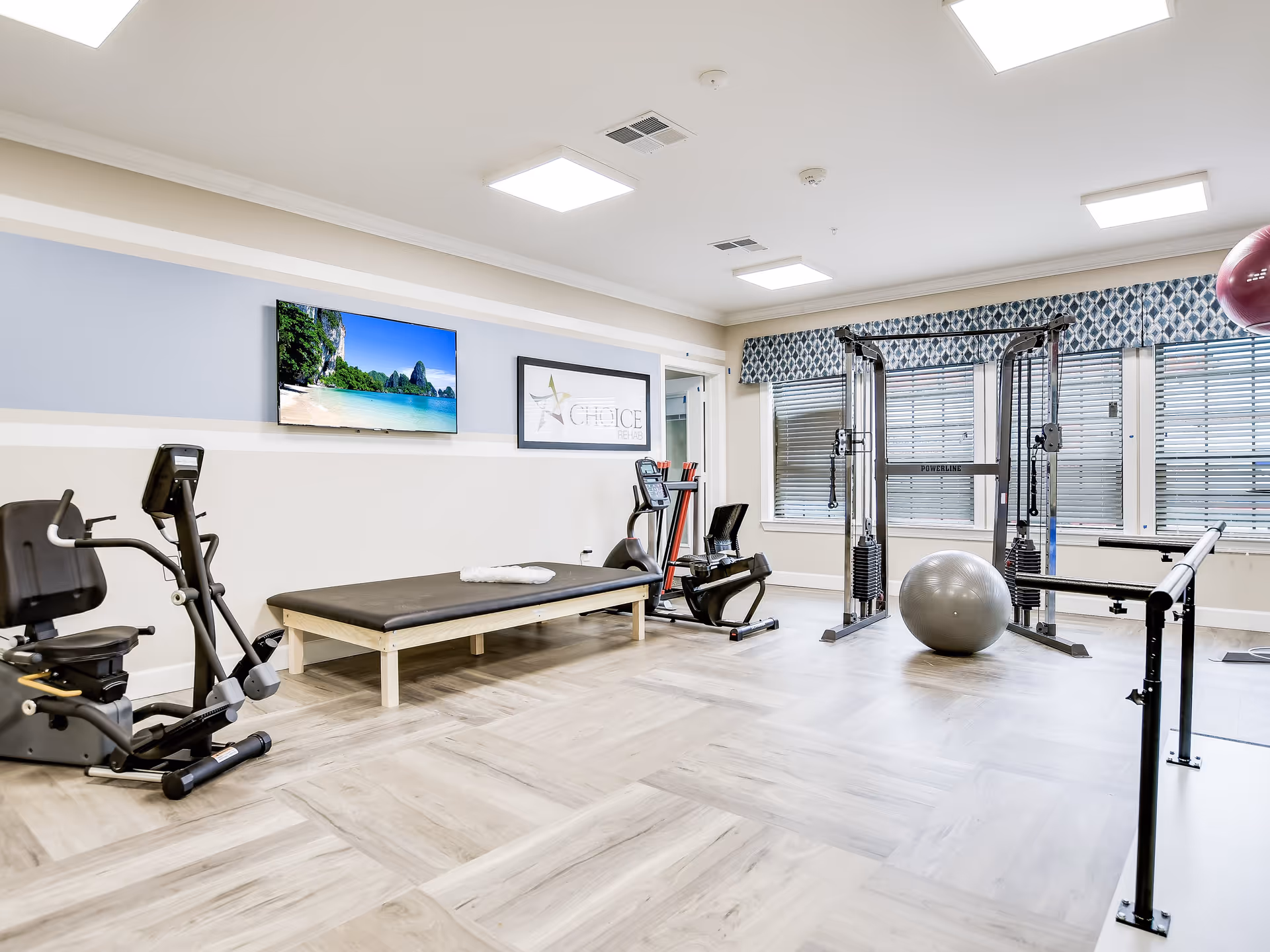 A bright and spacious fitness room in a senior living facility featuring exercise equipment including a recumbent bike, a treadmill, a multi-functional weight machine, a large exercise ball, and parallel bars. The room has light-colored wood flooring, large windows with patterned valances, and a wall-mounted TV displaying a beach scene.