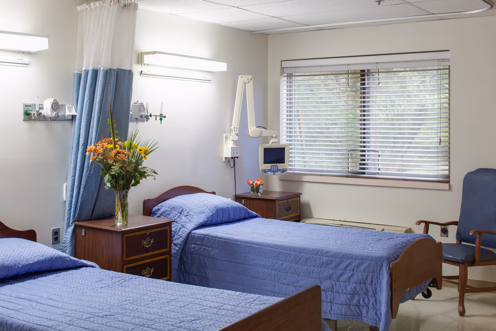 A room with two hospital-style beds covered in blue quilts, separated by a wooden nightstand with a vase of flowers. There is medical equipment mounted on the wall above the beds, a window with blinds, and a blue armchair in the corner.