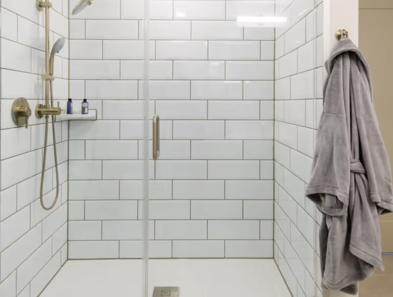Glass-enclosed shower with white subway tile, brass shower fixtures, and a gray bathrobe hanging on the right.