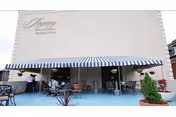 Outdoor patio area at Regency Healthcare & Rehab Center with several tables and chairs under blue and white striped awnings attached to the building. Potted plants and hanging flower baskets decorate the space.