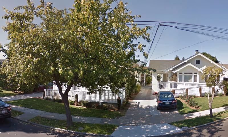 Exterior view of a single-story residential-style building with a white fence, a driveway with a parked car, and trees in the front yard under a clear sky.