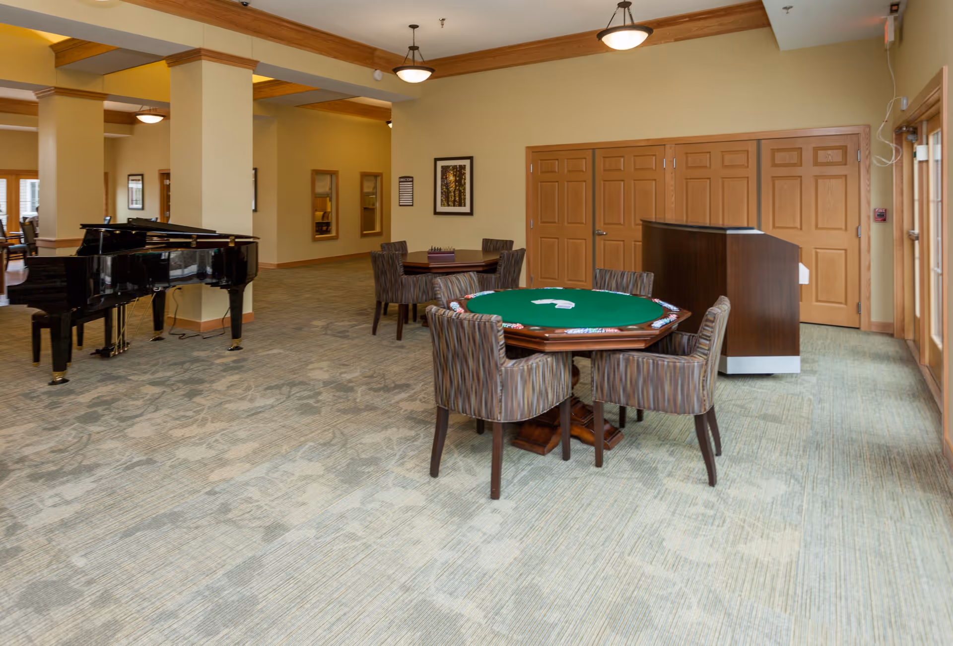 A spacious common area with a green felt card table surrounded by six patterned chairs, a black grand piano, and a wooden podium near a set of closed wooden double doors. The room has beige walls, carpeted floors with a subtle pattern, and ceiling lights.