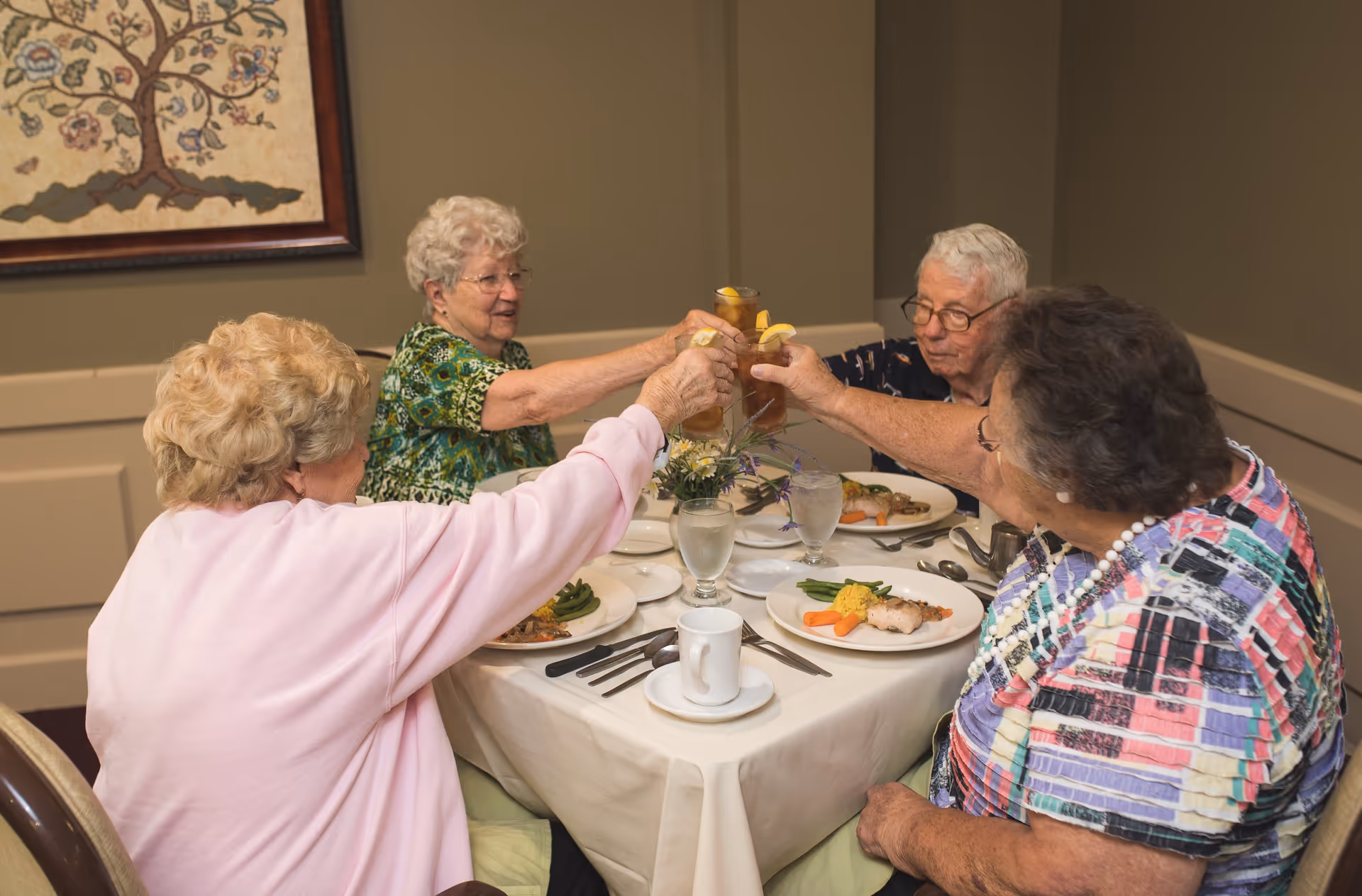 Four elderly women sitting around a dining table in a senior living facility, raising their glasses in a toast. The table is set with plates of food, glasses of water, and a small floral centerpiece. The room has neutral-colored walls and a framed artwork featuring a tree.