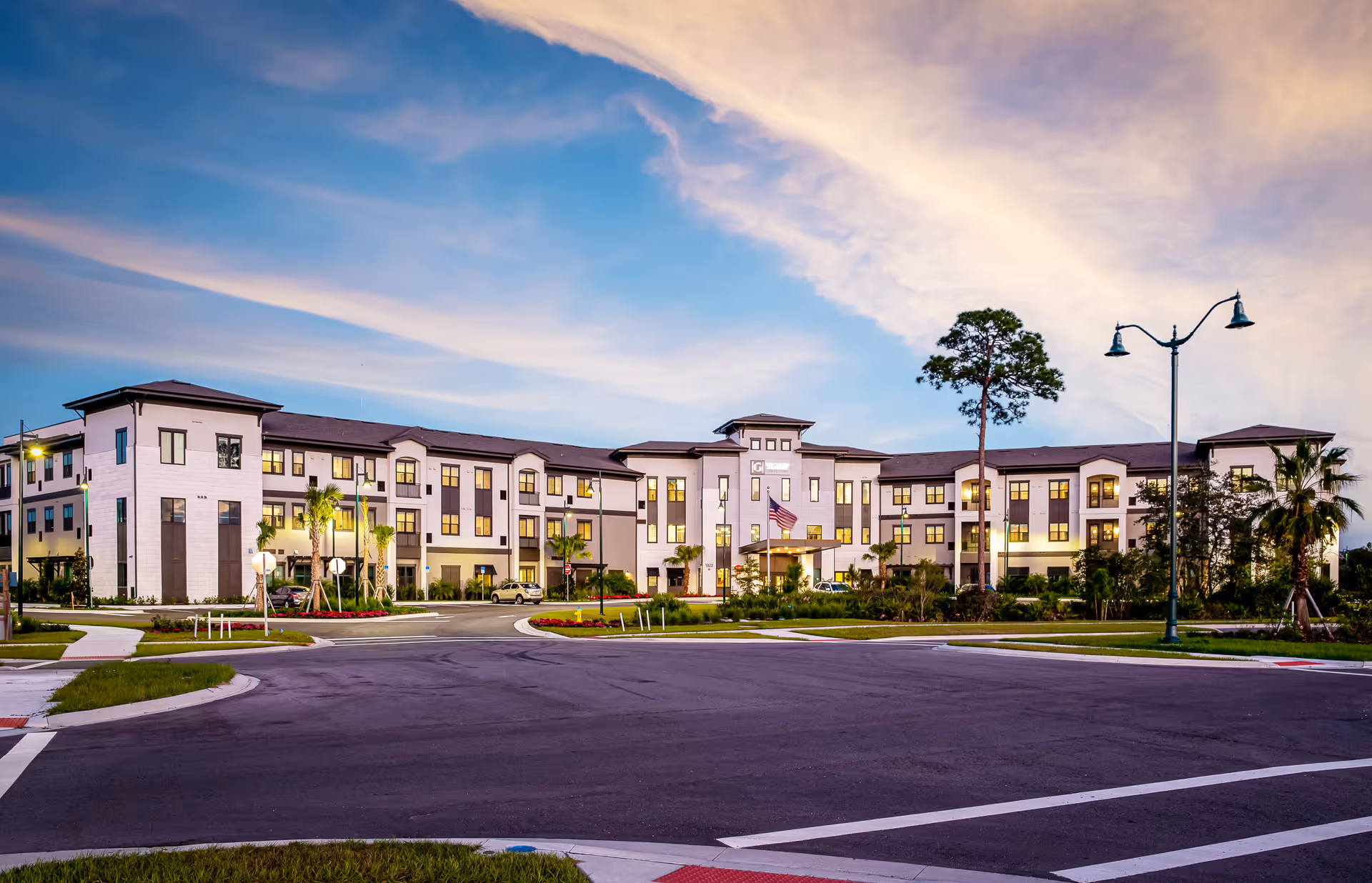 Front exterior of a three-story senior living facility with illuminated windows, landscaped entrance, and a dramatic evening sky.