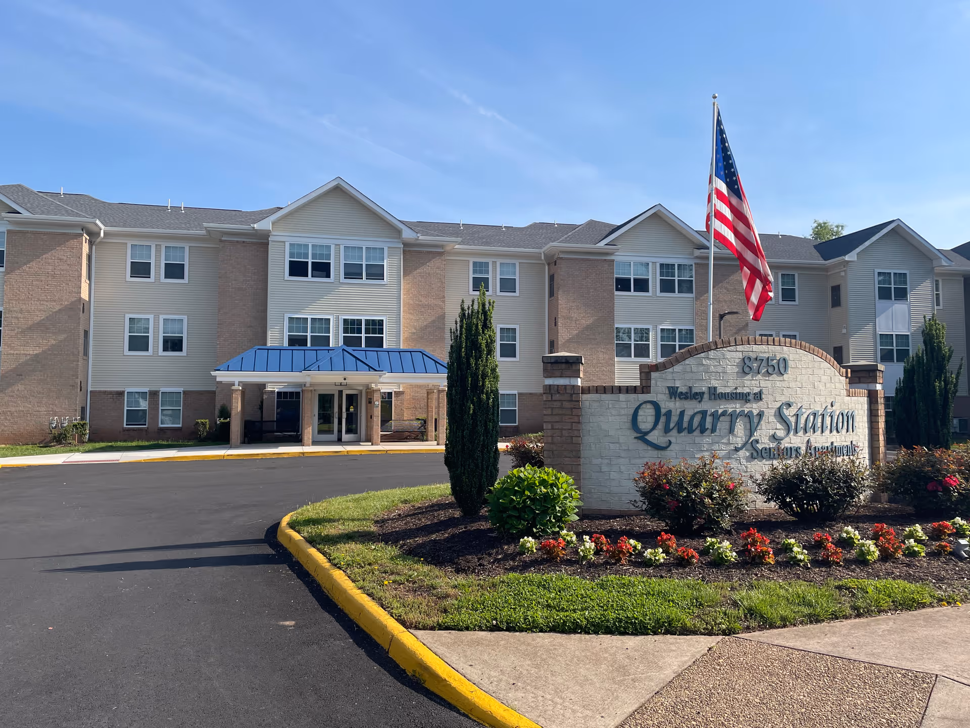 Exterior view of Quarry Station Seniors Apartments building with a three-story structure, beige siding, brick accents, and a blue awning over the entrance. In front of the building is a landscaped area with flowers, shrubs, a small tree, and a sign displaying the facility name and address. An American flag is flying on a pole behind the sign under a clear blue sky.