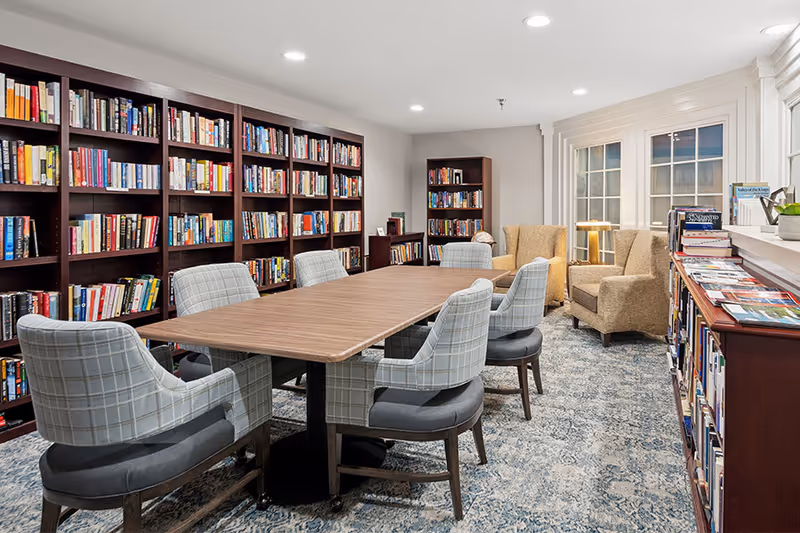 A cozy library room with a long wooden table surrounded by eight cushioned chairs. The walls are lined with dark wooden bookshelves filled with books. Two beige armchairs are placed near a window at the far end of the room. The floor is covered with a patterned carpet, and the room is well-lit with recessed ceiling lights.