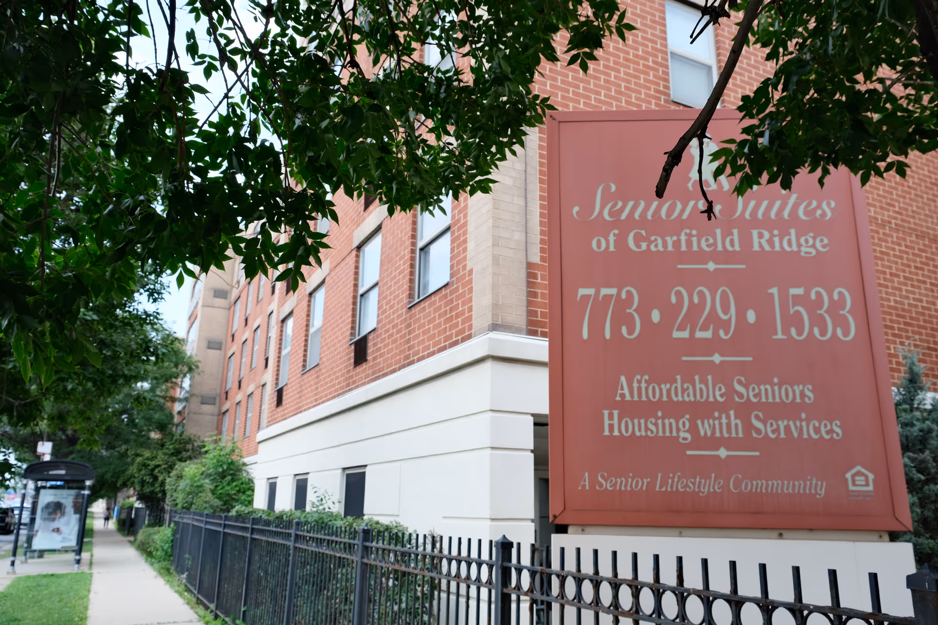 Exterior view of a brick senior living facility named Senior Suites of Garfield Ridge with a large red sign displaying the facility name, phone number, and description as affordable seniors housing with services. The building is partially obscured by green tree branches, and there is a black metal fence along the sidewalk in front.