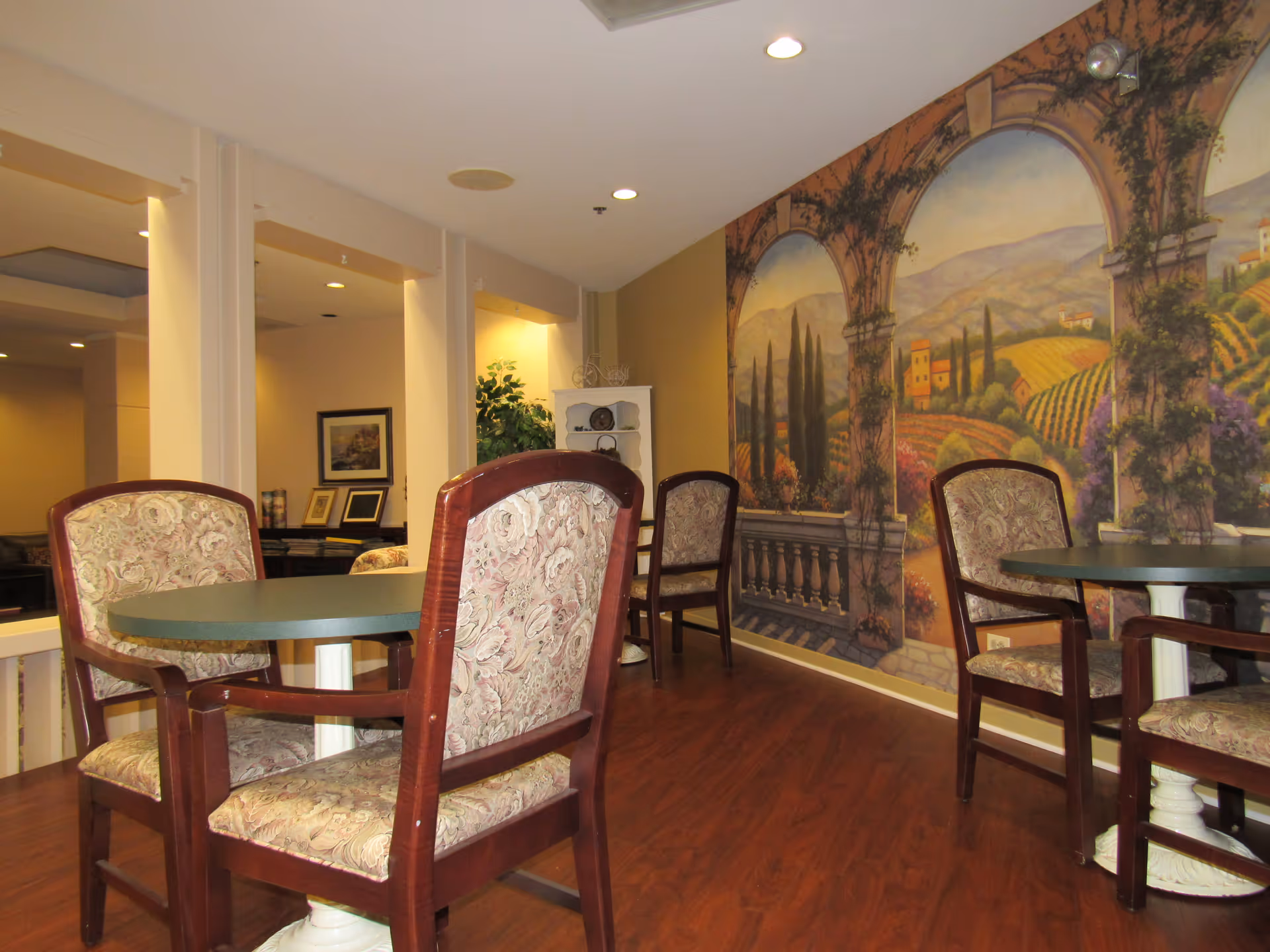 Small common dining area with round tables, upholstered chairs, and a large landscape mural on the wall.
