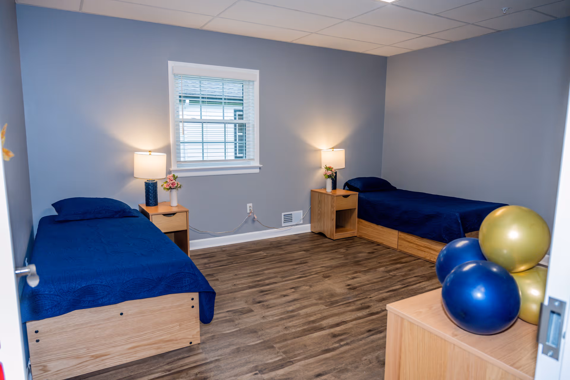 A simple bedroom with two single beds covered in dark blue bedding, each with a wooden nightstand holding a lamp and a small vase of flowers. The room has light gray walls, a window with white blinds, and a wooden floor. There are three balloons, two blue and one gold, on a wooden dresser near the door.