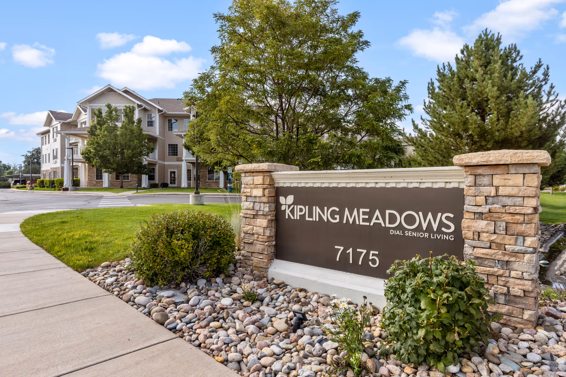 Stone sign reading 'Kipling Meadows' in front of a senior living building with trees, shrubs, and a sidewalk.