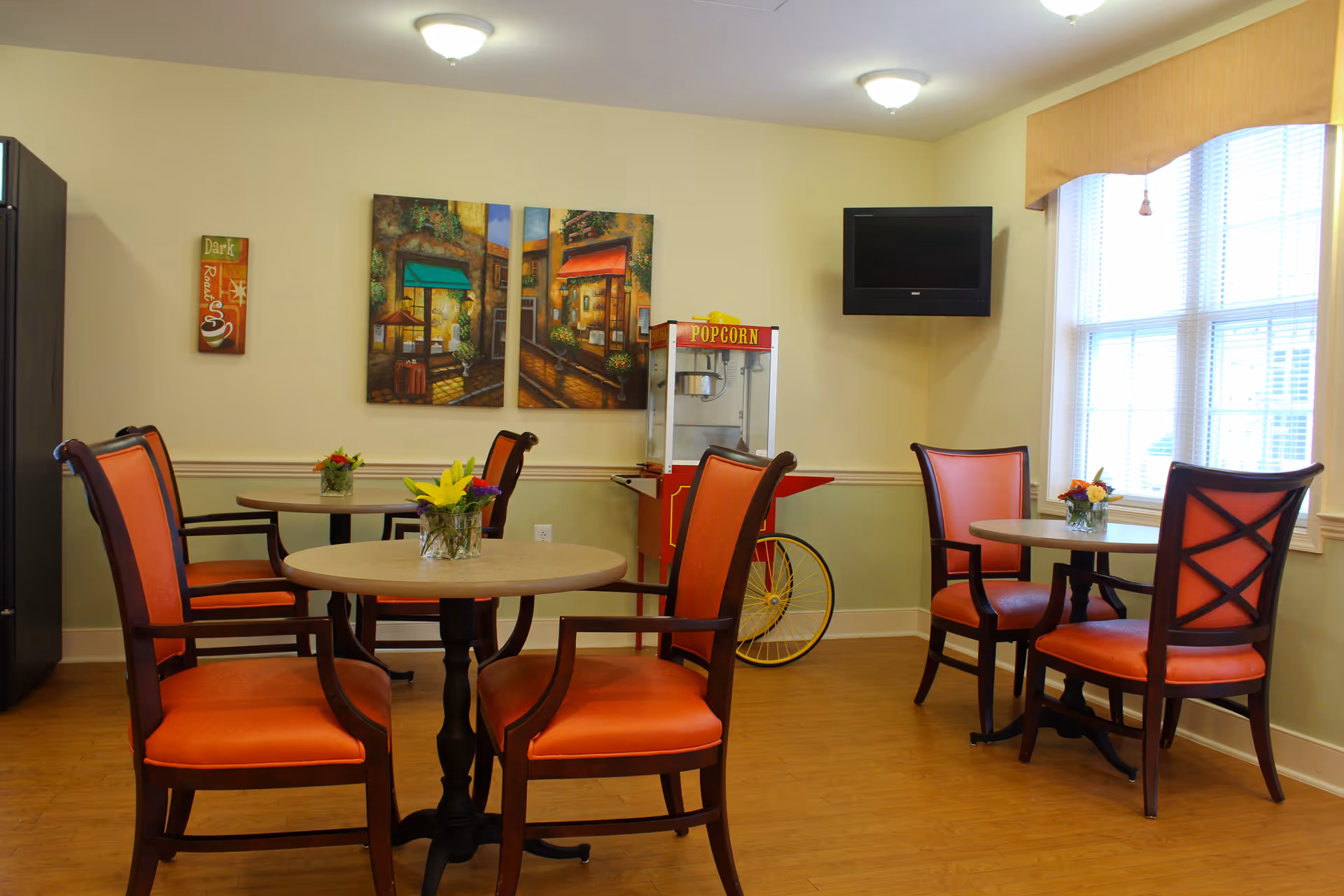 A cozy dining area with round tables and orange cushioned chairs. Each table has a small flower arrangement. There is a popcorn machine against the wall, a wall-mounted TV, and a large window letting in natural light. The walls are decorated with colorful paintings and a small coffee-themed wall art.