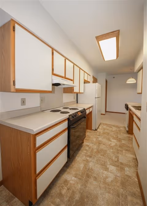 Galley-style kitchen with white cabinets trimmed in wood, an electric stove, refrigerator, and vinyl tile flooring.