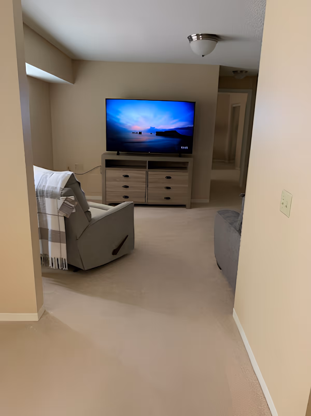 Small living room with a television on a dresser, a recliner draped with a blanket, and beige walls and carpet.