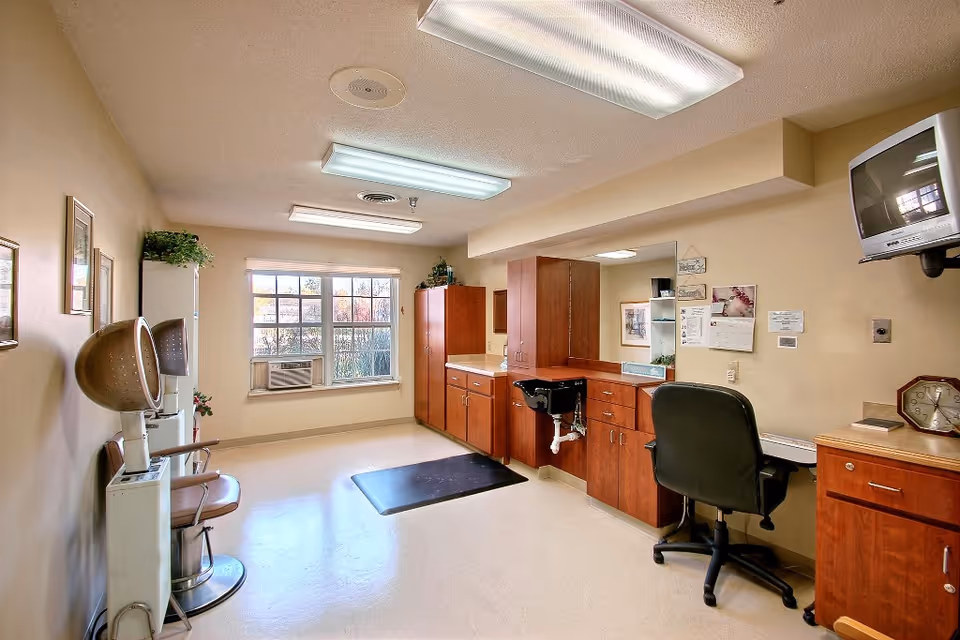 Interior view of a healthcare center room with beige walls and floor. The room features wooden cabinets, a black office chair in front of a counter with a mirror, a wall-mounted TV, and two vintage hair dryer chairs. A window with blinds lets in natural light, and fluorescent ceiling lights illuminate the space.