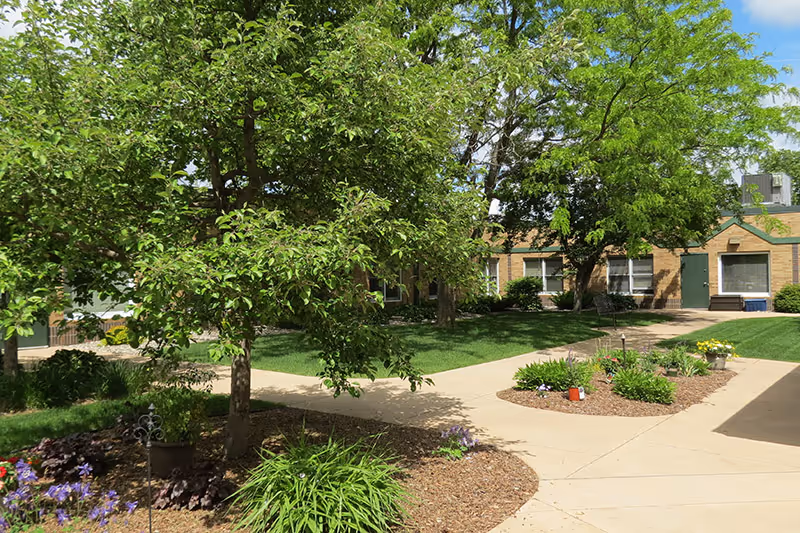 A sunny outdoor courtyard area with green trees, plants, and flower beds surrounded by a paved walkway. In the background, there is a single-story brick building with several windows and green doors.