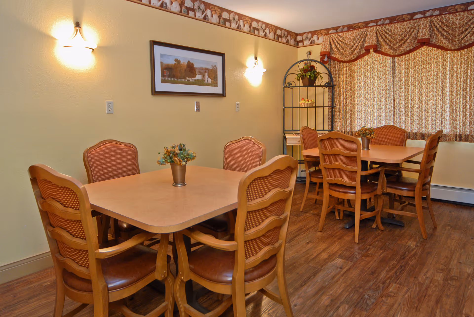 Dining room with two wooden tables and multiple matching chairs, wall sconces, framed artwork, shelving unit and patterned curtains.