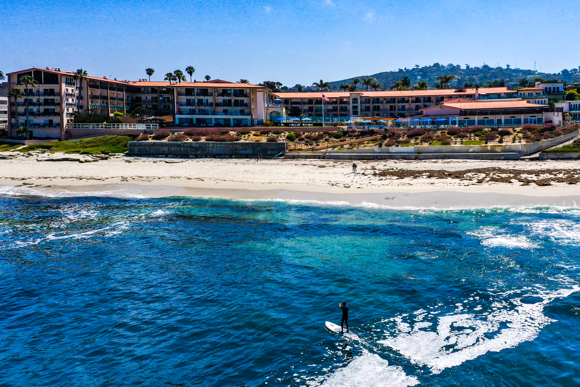A large multi-story building with balconies overlooking a sandy beach and blue ocean. A person is paddleboarding on the water near the shore. The building is surrounded by palm trees and has a clear blue sky above.