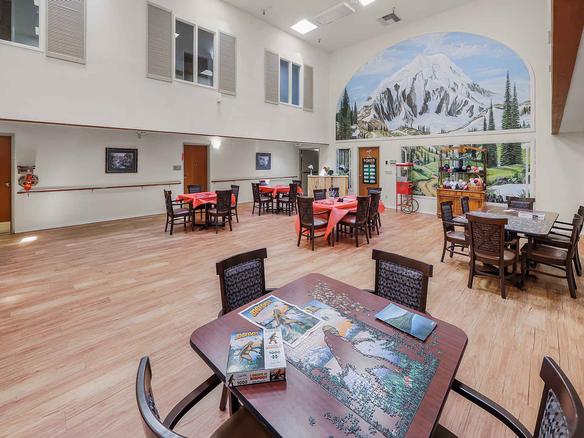 A spacious common area in an assisted living facility with several tables and chairs arranged for group activities. One table in the foreground has a partially completed jigsaw puzzle featuring a Bigfoot theme. The room has light wood flooring, high ceilings, and a large mural of a snowy mountain and forest on the far wall. There are windows high on the walls and various decorations including a popcorn machine and shelves with yarn balls.