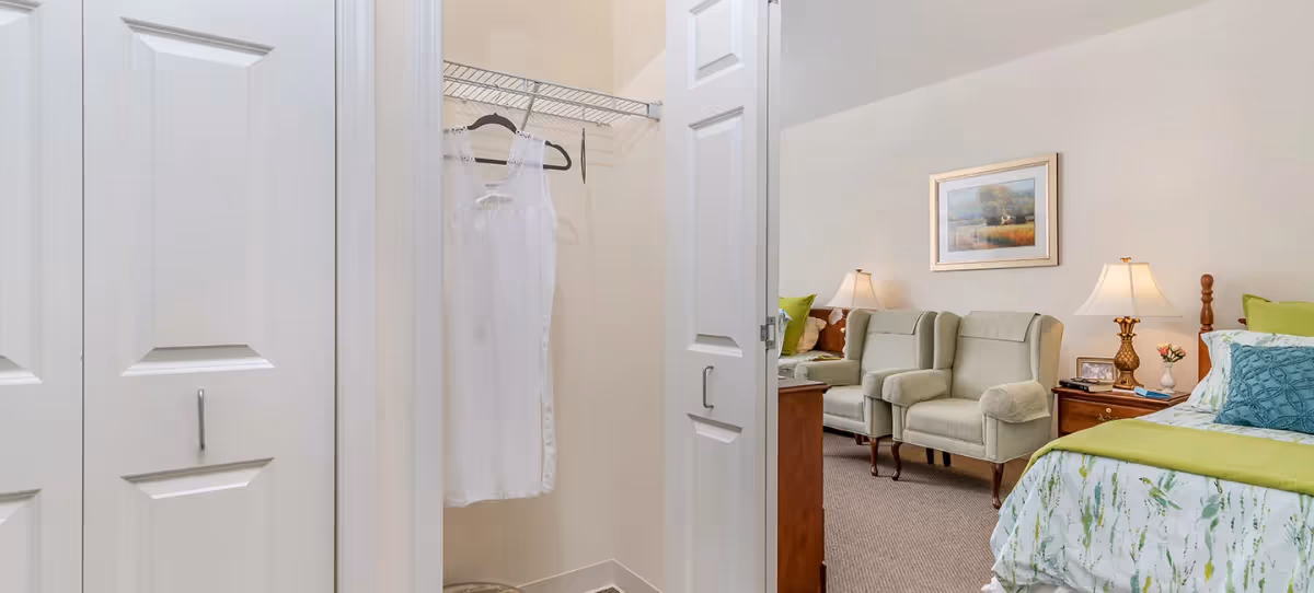 View of a bedroom with a bed covered in white and green bedding, two beige armchairs, a wooden nightstand with a lamp and flowers, and a framed painting on the wall. In the foreground, there is an open closet door revealing a white dress hanging inside.
