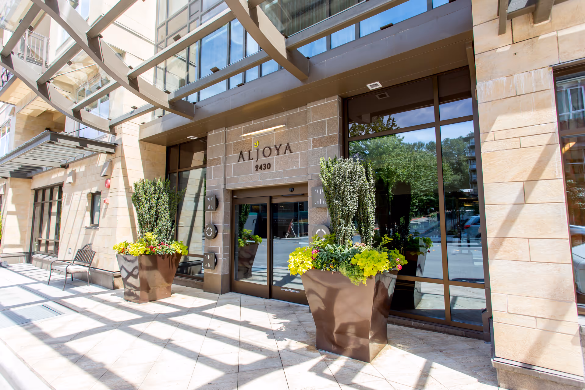 Entrance to Aljoya Mercer Island facility with large glass doors, two large planters filled with green and yellow plants, and a modern building facade with beige stone walls and glass windows.
