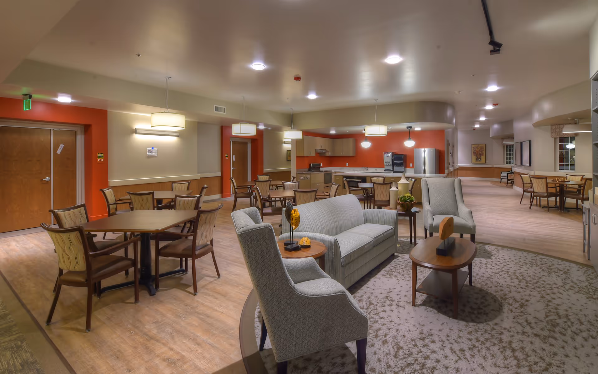 A spacious senior living community common area with multiple seating arrangements including a sofa, armchairs, and several dining tables with chairs. The room features warm lighting, wooden floors, and a kitchen area with cabinets, a refrigerator, and a coffee machine in the background. The walls are painted in neutral tones with some orange accent walls.